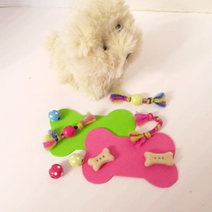May include: A white plush toy dog with two green and pink bone-shaped toys. The toys have colourful strings and beads attached to them.