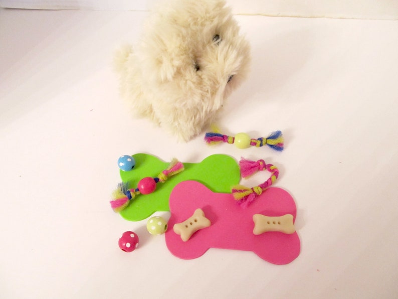 May include: A white plush toy dog with two green and pink bone-shaped toys. The toys have colourful strings and beads attached to them.