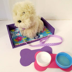 May include: A white plush dog toy in a purple bed with a floral patterned blue blanket. The dog toy has a purple leash with white polka dots. There are two purple bone-shaped mats with a pink bone-shaped mat in the middle. The pink mat has two white and blue bowls on top.