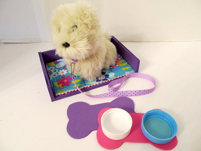 May include: A white plush dog toy in a purple bed with a floral patterned blue blanket. The dog toy has a purple leash with white polka dots. There are two purple bone-shaped mats with a pink bone-shaped mat in the middle. The pink mat has two white and blue bowls on top.