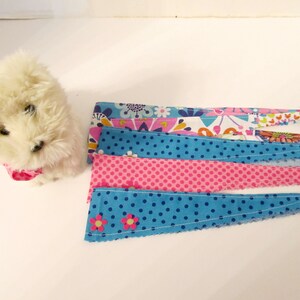 May include: A white plush toy dog wearing a pink fabric collar. The dog is sitting on a white surface. There are five fabric strips with different patterns, including floral, polka dots, and solid colours, arranged in a row below the dog.