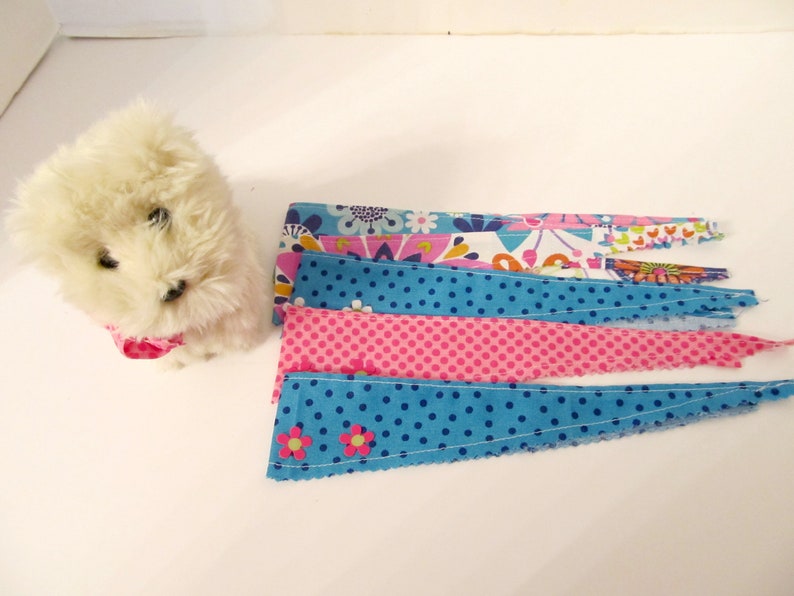 May include: A white plush toy dog wearing a pink fabric collar. The dog is sitting on a white surface. There are five fabric strips with different patterns, including floral, polka dots, and solid colours, arranged in a row below the dog.