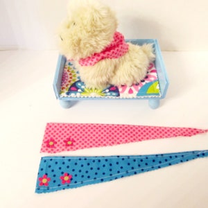 May include: Two fabric scraps with pink and blue polka dots and pink flowers. The scraps are triangular in shape and have a raw edge. A small stuffed white dog is sitting on a blue bed with a colourful patterned mattress.