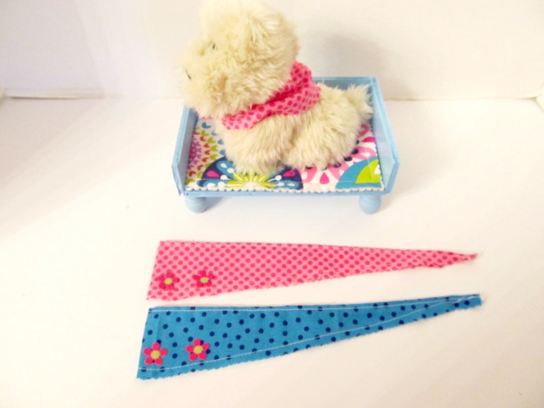 May include: Two fabric scraps with pink and blue polka dots and pink flowers. The scraps are triangular in shape and have a raw edge. A small stuffed white dog is sitting on a blue bed with a colourful patterned mattress.