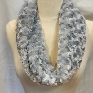 May include: A light gray infinity scarf made of soft, faux fur. The scarf is displayed on a mannequin and has a textured, rose-like pattern. The scarf is a winter accessory.