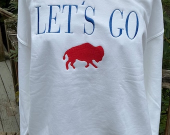 Könnte beinhalten: Ein weißes Sweatshirt mit Rundhalsausschnitt und dem gestickten Text LET'S GO in blauen Großbuchstaben. Unter dem Text befindet sich eine gestickte rote Silhouette eines Büffels. Das Kleidungsstück hat lange Ärmel sowie gerippte Bündchen.