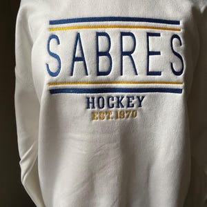 May include: White sweatshirt with the word "SABRES" embroidered in blue, with a gold and blue horizontal stripe above and below the word. Below the word "SABRES" is the word "HOCKEY" and the text "EST.1970" in gold.