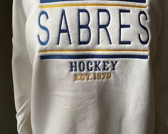 Könnte beinhalten: Weißes Sweatshirt mit dem Wort "SABRES" in Blau gestickt, mit einem goldenen und blauen horizontalen Streifen darüber und darunter. Unter dem Wort "SABRES" stehen die Wörter "HOCKEY" und der Text "EST.1970" in Gold.