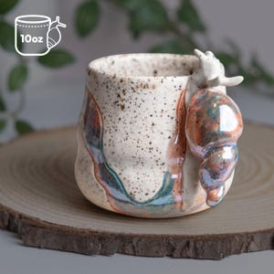 May include: A handmade ceramic mug with a snail design. The mug is off-white with brown speckles and colorful iridescent accents. The snail's shell is orange and blue. The mug holds 10 oz.