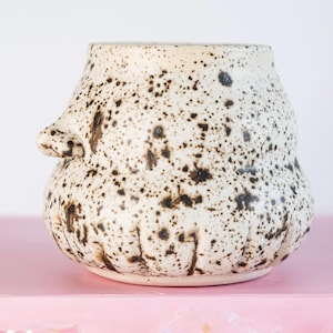 May include: A speckled ceramic pot with a cream-colored base and dark brown spots. The pot has a rounded body with a small handle on one side. The pot is sitting on a pink surface.