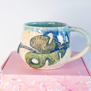 May include: A ceramic mug with a large handle, featuring a hand-painted design of a green and blue leaf. The mug has a speckled beige body with a blue rim and accents. The mug is sitting on a pink box.