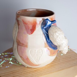 May include: A handmade ceramic mug with a seahorse-shaped handle. The mug features a white base with pink and orange accents, a blue seahorse handle, and a textured white element. A heart is carved into the side.