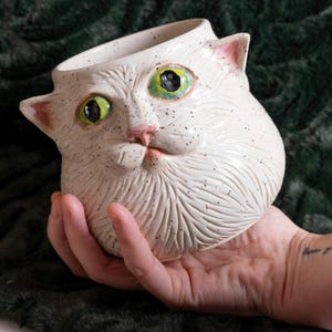 May include: A white ceramic cat-shaped mug with speckled details. The mug features a cat face with green and black eyes, a pink nose, and pink inner ears. The mug is held in a hand.