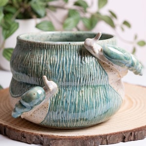 May include: A handcrafted ceramic planter with a textured, teal and beige exterior. Two snail sculptures are attached to the planter's side. The planter sits on a round wooden slice, showcasing a unique, nature-inspired design.
