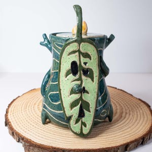 May include: A handcrafted ceramic jar with a lid, featuring a unique leaf-shaped handle and a decorative leaf design on the front. The jar is primarily teal with green and white accents, sitting on a wooden slice.