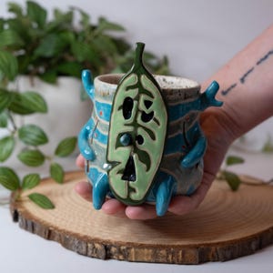 May include: A whimsical ceramic mug with a unique design. The mug is primarily blue with turquoise accents and a green leaf-shaped face. The mug has a textured surface and is held on a wooden slice.