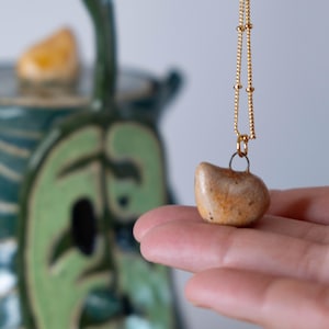 May include: A small, heart-shaped pendant necklace with a gold chain. The pendant is a natural stone with tan and orange hues. The necklace is displayed against a blurred background featuring a green and blue ceramic piece.