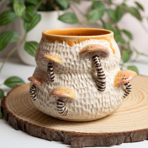 May include: A cream-colored ceramic planter with a textured surface and an orange rim. The planter is adorned with several mushroom-shaped decorations with brown and tan caps and striped stems. The planter sits on a wooden slice.