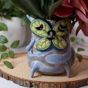 May include: A whimsical ceramic vase with a butterfly design, featuring green, yellow, and black accents. The vase has a blue and gray color scheme and is filled with flowers. It is placed on a wooden slice.