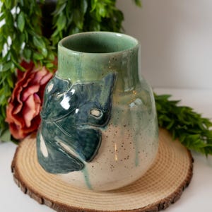 May include: A ceramic vase with a green and white glaze. The vase has a leaf design on the front.