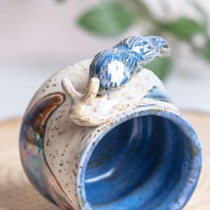 May include: A ceramic mug with a whimsical snail design. The mug features a speckled cream and brown exterior with a vibrant blue interior. A detailed blue snail sculpture adorns the rim, adding a unique, artistic touch to this handmade drinkware.