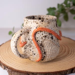 May include: A ceramic mug with a speckled cream-colored glaze featuring a curved orange clay accent wrapped around the exterior. The surface includes a textured, carved pattern on one side and a sturdy handle, creating a unique tactile appearance.