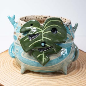 May include: A handmade ceramic planter with a whimsical design. The planter is primarily turquoise with a green leaf accent and a speckled rim. It features three small feet and two curved handles, perfect for small plants.