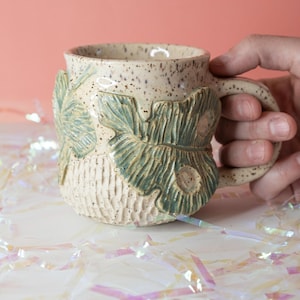 May include: A ceramic mug with a speckled cream finish, featuring raised green leaf motifs on the exterior. The lower portion of the mug has a carved, textured pattern, and it includes a sturdy handle on the side for a comfortable grip.