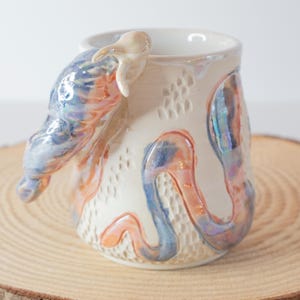 May include: A ceramic mug with a cream-colored base featuring an iridescent, swirling blue and orange sculptural handle and decorative accents. The surface has a textured, dimpled pattern across the body, with a glossy, reflective finish.