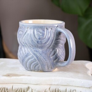 May include: A handmade ceramic mug with a blue and white glaze. The mug features a textured, carved design and a rounded handle. The interior is a cream color. This unique mug is perfect for enjoying coffee, tea, or other beverages.