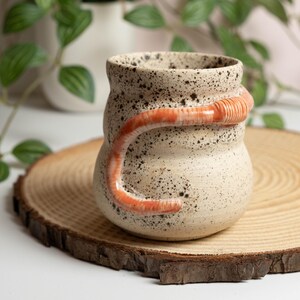 May include: A speckled ceramic mug with a curved, orange handle. The mug is off-white with dark brown speckles. The handle is a vibrant orange with a textured design. The mug sits on a round wooden slice.