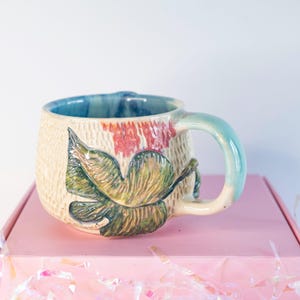 May include: Handmade ceramic mug with a textured beige exterior and a blue interior. The mug features a green and yellow leaf design and a light blue handle. Red floral accents are visible near the rim. The mug is sitting on a pink box.