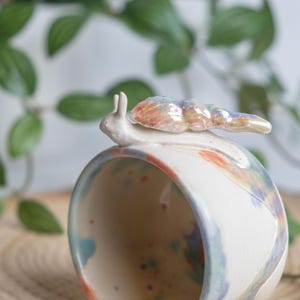 May include: A ceramic mug with a whimsical snail figurine on top. The mug is white with colorful paint splatters in shades of blue, orange, and red. The snail has a pearlescent shell. The mug is a unique piece of home decor.