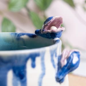 May include: A handmade ceramic mug with a unique design. The mug features a blue and white exterior with a pearl-like bead inside a shell-shaped handle. The shell is blue with a pink interior.