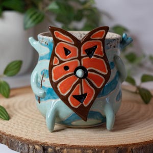May include: A whimsical ceramic planter with a light blue base and a brown and orange owl-like face. The planter has blue stripes and small arms and legs. The planter is sitting on a wooden surface.