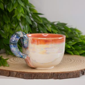 May include: A ceramic coffee mug with a unique, iridescent glaze. The mug has a rounded shape and a handle that is a different color than the mug itself. The handle is a swirling blue and green color.