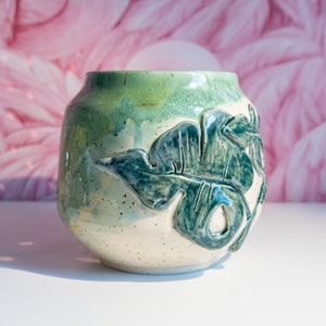 May include: A white ceramic planter with a green glaze and a raised leaf design. The planter has a speckled finish and is perfect for adding a touch of nature to any space.