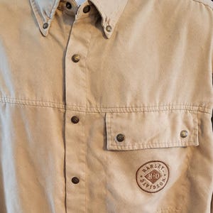 Może przedstawiać: Beżowa koszula zapinana na guziki z długimi rękawami. Posiada kołnierzyk zapinany na guziki, kieszeń na piersi i naszywkę z napisem "HARLEY-DAVIDSON H-D". Koszula ma ciemnobrązowe guziki i przeszycia.