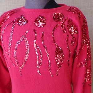 Pode incluir: Um suéter vermelho brilhante com mangas compridas e gola redonda. O suéter é adornado com lantejoulas vermelhas em um padrão floral no peito e nos ombros. Os punhos e a bainha são canelados.