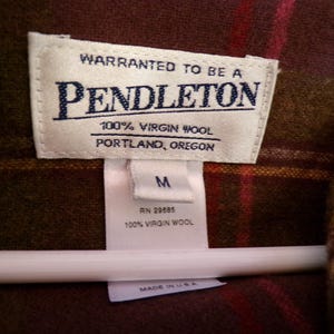 Puede incluir: Primer plano de una etiqueta Pendleton sobre una tela a cuadros marr&oacute;n y roja. La etiqueta dice "WARRANTED TO BE A PENDLETON 100% VIRGIN WOOL PORTLAND, OREGON". Debajo de la etiqueta hay una etiqueta m&aacute;s peque&ntilde;a con la talla "M" y "100% VIRGIN WOOL".