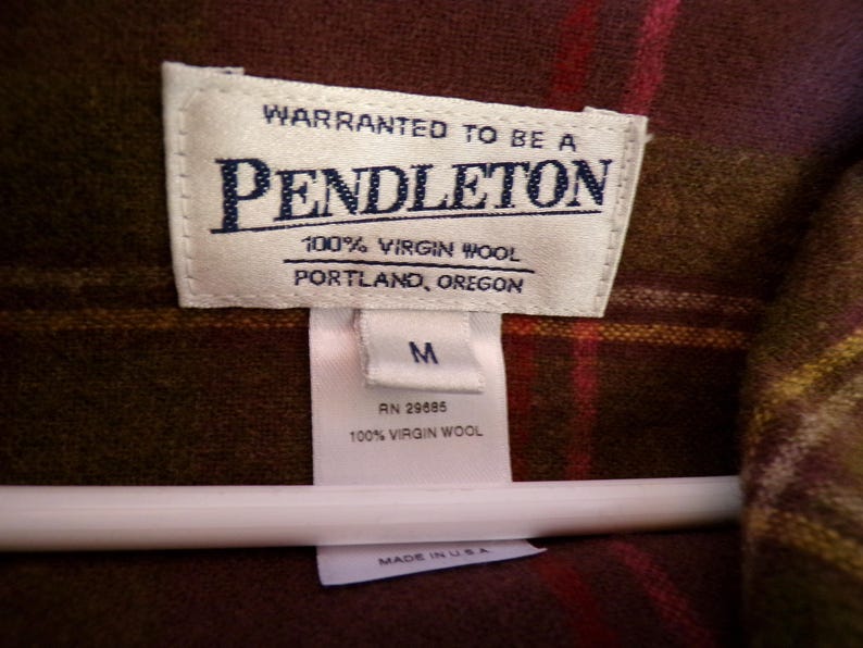 Puede incluir: Primer plano de una etiqueta Pendleton sobre una tela a cuadros marr&oacute;n y roja. La etiqueta dice "WARRANTED TO BE A PENDLETON 100% VIRGIN WOOL PORTLAND, OREGON". Debajo de la etiqueta hay una etiqueta m&aacute;s peque&ntilde;a con la talla "M" y "100% VIRGIN WOOL".