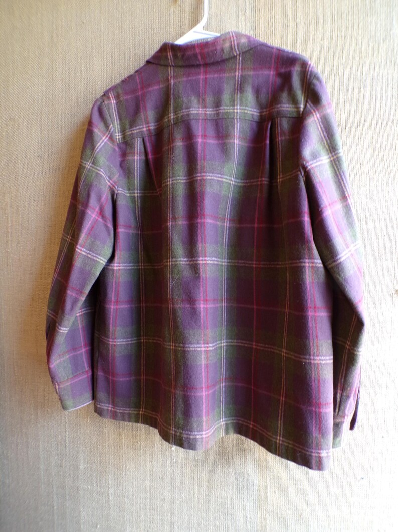 Puede incluir: Una camisa chaqueta a cuadros morados, verdes y rosas cuelga de una percha blanca. La parte trasera de la chaqueta es visible, mostrando el cuello, los pu&ntilde;os abotonados y un canes&uacute; plisado en la espalda. El estampado de cuadros es un dise&ntilde;o cl&aacute;sico.