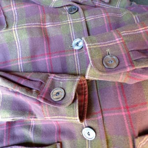 Puede incluir: Primer plano de una chaqueta de cuadros Pendleton en tonos morado, verde y rojo. La chaqueta presenta una botonadura frontal, pu&ntilde;os abotonados y un cuello. La tela tiene un patr&oacute;n de cuadros cl&aacute;sico.