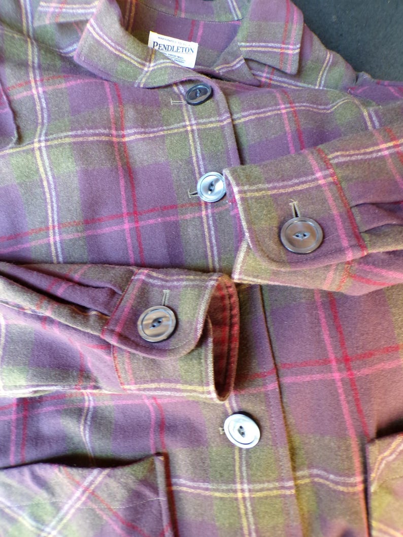 Puede incluir: Primer plano de una chaqueta de cuadros Pendleton en tonos morado, verde y rojo. La chaqueta presenta una botonadura frontal, pu&ntilde;os abotonados y un cuello. La tela tiene un patr&oacute;n de cuadros cl&aacute;sico.