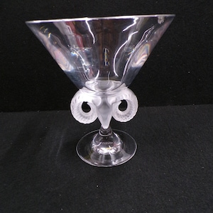 May include: A clear glass martini glass with a wide, triangular bowl and a decorative base. The base features a frosted design with two curved elements. The glass sits on a round, clear base.