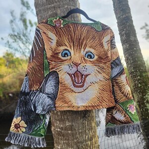 May include: A tapestry-style cropped sweater featuring a large, smiling orange tabby cat face. The sleeves showcase a gray cat and floral accents, with fringed edges. The sweater is hanging on a black hanger against a natural background.
