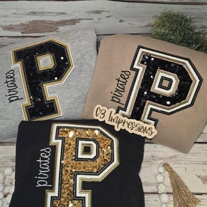 May include: Three sweatshirts with a large letter "P" applique. The "P" is black or gold with sequins, outlined in white and gold, and the word "pirates" is embroidered on the side. One sweatshirt is gray, one is tan, and one is black.
