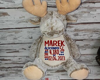 May include: A plush moose toy with tan antlers and cream-colored accents. The moose has embroidered text on its chest that reads "MAREK", "ALSKANDER 56", "4.180 cm", and "02.06.2023". The moose is sitting on a white wooden surface.
