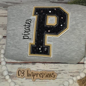 May include: A gray sweatshirt with a large black sequined letter "P" outlined in gold and white. The word "pirates" is written in black script on the left side. The sweatshirt has a crew neck and is likely made of a soft material.