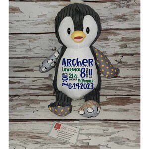 May include: A gray and white penguin plush toy with embroidered details. The penguin has yellow polka dots on one wing and a white belly with the text "Archer Lawrence 8:01 AM 21 1/2 inches 8 oz McDonald 6-29-2023" embroidered on it.